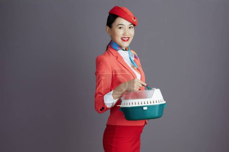 Photo for Happy stylish asian female flight attendant in red skirt, jacket and hat uniform with pet carrier isolated on gray background. - Royalty Free Image