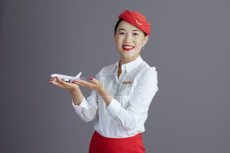 Photo for Smiling modern flight attendant asian woman in red skirt and hat uniform with a little airplane against gray background. - Royalty Free Image