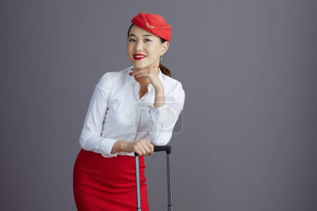 Foto de Feliz moderno asiático femenino vuelo asistente en rojo falda y sombrero uniforme con rueda bolsa aislado en gris fondo. - Imagen libre de derechos