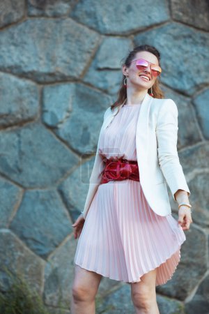 Téléchargez les photos : Heureuse femme élégante de 40 ans en robe rose et veste blanche dans la ville avec des lunettes de soleil contre le mur de pierre. - en image libre de droit