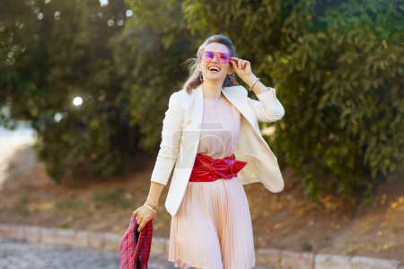 Foto de Mujer moderna feliz en vestido rosa y chaqueta blanca en la ciudad con bolso rojo y gafas de sol caminando. - Imagen libre de derechos