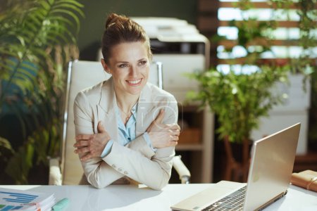 Foto de Feliz mujer trabajadora moderna en un traje de negocios ligero en la oficina verde moderna con el ordenador portátil abrazándose. - Imagen libre de derechos