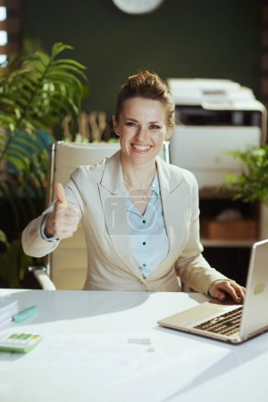 Foto de Retrato de mujer trabajadora moderna sonriente en un traje de negocios ligero en la oficina verde moderna con el ordenador portátil que muestra los pulgares hacia arriba. - Imagen libre de derechos