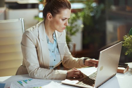 Téléchargez les photos : Femme comptable d'âge moyen moderne dans un costume d'affaires léger dans un bureau vert moderne avec ordinateur portable. - en image libre de droit