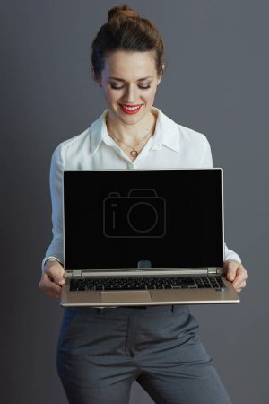 Foto de Sonriente joven dueño de una pequeña empresa mujer en blusa blanca que muestra la pantalla en blanco portátil aislado en fondo gris. - Imagen libre de derechos