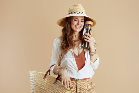 Foto de Vacaciones en playa. sonriente mujer de mediana edad de moda en blusa blanca y pantalones cortos contra fondo beige con botella de agua, bolsa de paja y sombrero de paja. - Imagen libre de derechos