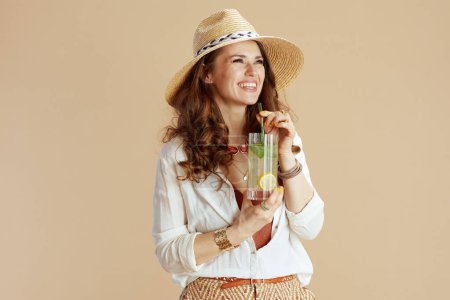 Foto de Vacaciones en playa. Sonriente mujer moderna en blusa blanca y pantalones cortos sobre fondo beige con cóctel y sombrero de verano. - Imagen libre de derechos