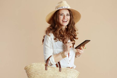 Téléchargez les photos : Vacances à la plage. heureuse femme élégante en chemisier blanc et short sur fond beige avec sac de paille et chapeau d'été en utilisant smartphone. - en image libre de droit