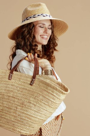 Photo for Beach vacation. smiling trendy 40 years old woman in white blouse and shorts isolated on beige background with straw bag and straw hat. - Royalty Free Image