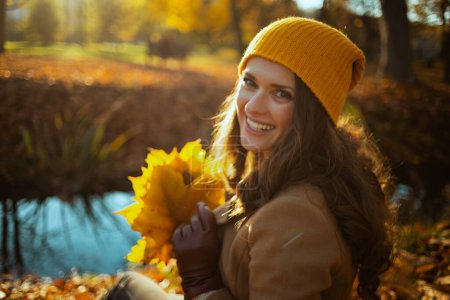 Téléchargez les photos : Bonjour, novembre. heureuse femme à la mode de 40 ans en manteau marron et chapeau jaune avec des feuilles jaunes d'automne assis à l'extérieur dans le parc de la ville en automne. - en image libre de droit