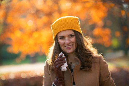 Téléchargez les photos : Bonjour septembre. Femme élégante et souriante en manteau beige et chapeau orange avec serviette mouchant à l'extérieur sur le parc de la ville en automne. - en image libre de droit