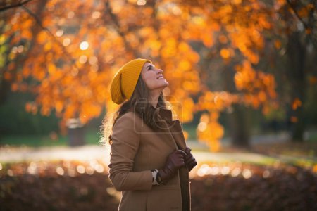 Hello october. smiling stylish female in beige coat and orange hat outdoors on the city park in autumn.