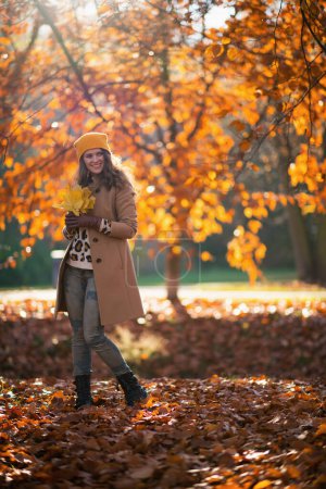 Photo for Hello november. Full length portrait of smiling stylish 40 years old woman in beige coat and orange hat with autumn yellow leaves outdoors on the city park in autumn. - Royalty Free Image
