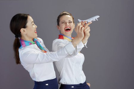 Photo for Happy elegant air hostess women in blue skirt, white shirt and scarf with a little airplane isolated on grey background. - Royalty Free Image