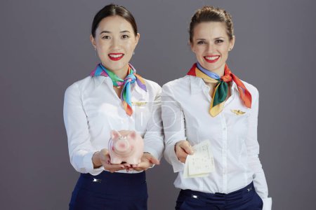 Photo for Smiling stylish air hostess women in blue skirt, white shirt and scarf with flight tickets and piggy bank isolated on grey background. - Royalty Free Image