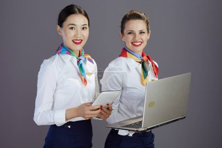 Photo for Smiling modern air hostess women in blue skirt, white shirt and scarf with tablet PC using laptop isolated on gray background. - Royalty Free Image