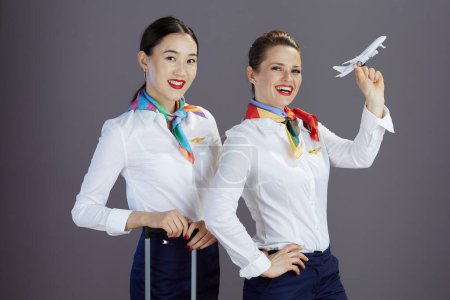 Téléchargez les photos : Joyeuses hôtesses de l'air féminines élégantes en jupe bleue, chemise blanche et écharpe avec un petit avion et sac trolley isolé sur fond gris. - en image libre de droit