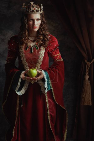 Foto de Reina medieval en vestido rojo con manzana verde y corona sobre fondo gris oscuro. - Imagen libre de derechos