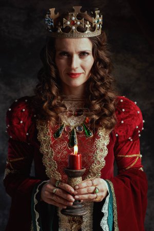 Foto de Reina medieval sonriente en vestido rojo con vela y corona sobre fondo gris oscuro. - Imagen libre de derechos