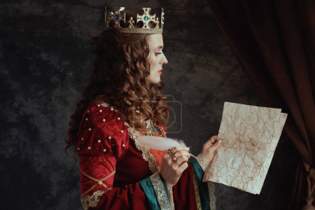 Photo for Medieval queen in red dress with parchment and crown on dark gray background. - Royalty Free Image