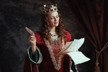 Foto de Feliz reina medieval en vestido rojo con pergamino y corona sobre fondo gris oscuro. - Imagen libre de derechos