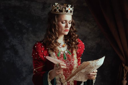 Photo for Pensive medieval queen in red dress with parchment and crown on dark gray background. - Royalty Free Image