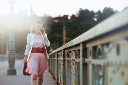 Photo for Happy stylish 40 years old woman in pink dress and white jacket in the city speaking on a smartphone on the bridge. - Royalty Free Image