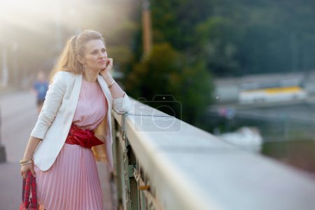 Photo for Pensive elegant woman in pink dress and white jacket in the city on the bridge. - Royalty Free Image