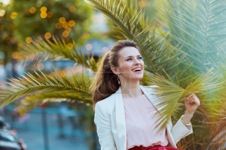 Téléchargez les photos : Femme élégante et souriante en robe rose et veste blanche dans la ville près d'un arbre tropical. - en image libre de droit