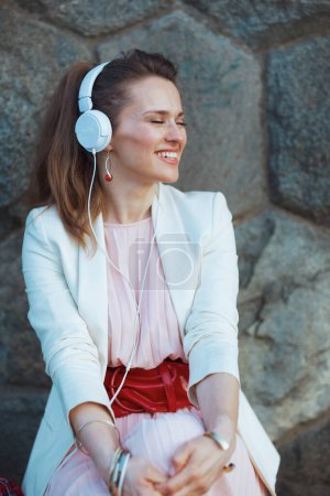 Téléchargez les photos : Heureuse femme à la mode en robe rose et veste blanche dans la ville avec écouteurs contre mur de pierre. - en image libre de droit