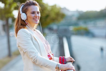 Téléchargez les photos : Femme souriante à la mode en robe rose et veste blanche dans la ville avec écouteurs. - en image libre de droit
