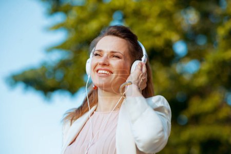 Foto de Sonriente elegante mujer de 40 años en vestido rosa y chaqueta blanca en la ciudad escuchando la música con auriculares. - Imagen libre de derechos