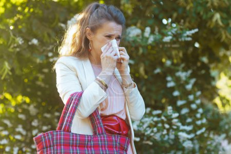 Téléchargez les photos : Tendance 40 ans femme en robe rose et veste blanche dans la ville avec serviette et sac rouge a une attaque d'allergie. - en image libre de droit