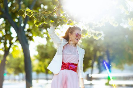 Foto de Mujer moderna feliz en vestido rosa y chaqueta blanca en la ciudad con gafas de sol. - Imagen libre de derechos