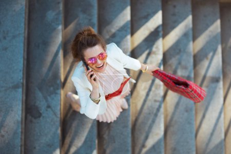 Foto de Vista superior de mujer elegante sonriente en vestido rosa y chaqueta blanca en la ciudad con bolsa roja y gafas de sol usando un teléfono inteligente. - Imagen libre de derechos