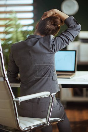 Sustainable workplace. Seen from behind modern small business owner woman in a grey business suit in modern green office stretching neck.