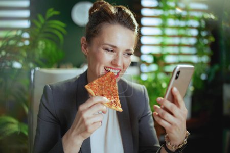 Photo for Sustainable workplace. smiling modern middle aged woman worker in a grey business suit in modern green office with pizza using smartphone applications. - Royalty Free Image