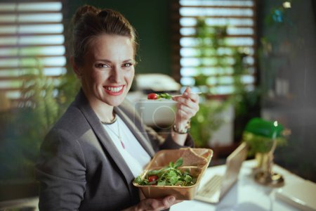 Sustainable workplace. happy modern 40 years old bookkeeper woman in a grey business suit in modern green office with laptop eating salad.