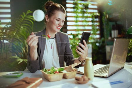 Téléchargez les photos : Un lieu de travail durable. travailleuse moderne souriante dans un costume d'affaires gris dans un bureau vert moderne avec ordinateur portable et smartphone manger de la salade. - en image libre de droit