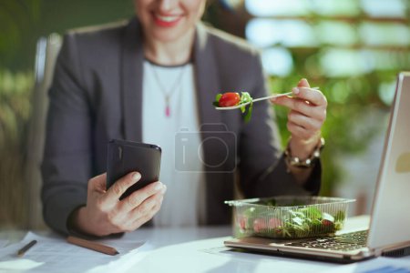 Téléchargez les photos : Un lieu de travail durable. heureuse femme d'affaires moderne dans un costume d'affaires gris dans un bureau vert moderne avec ordinateur portable et smartphone manger de la salade. - en image libre de droit