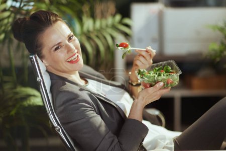 Photo for Sustainable workplace. smiling modern bookkeeper woman in a grey business suit in modern green office eating salad. - Royalty Free Image