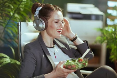 Photo for Sustainable workplace. happy modern middle aged accountant woman in a grey business suit in modern green office with headphones eating salad. - Royalty Free Image