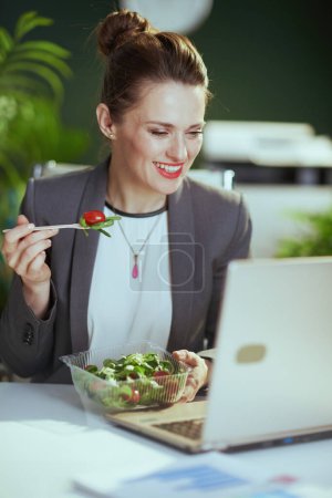 Foto de Lugar de trabajo sostenible. contable moderna feliz mujer en un traje de negocios gris en la oficina verde moderna con portátil comer ensalada. - Imagen libre de derechos