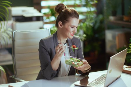 Téléchargez les photos : Un lieu de travail durable. souriant femme d'âge moyen moderne travailleur dans un costume d'affaires gris dans un bureau vert moderne avec ordinateur portable manger salade. - en image libre de droit