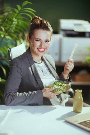 Téléchargez les photos : Un lieu de travail durable. heureuse employée moderne dans un costume d'affaires gris dans un bureau vert moderne mangeant de la salade. - en image libre de droit