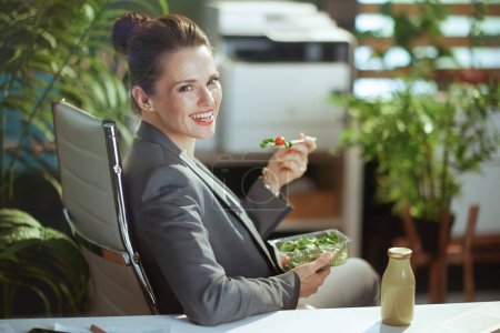 Téléchargez les photos : Un lieu de travail durable. heureuse ouvrière moderne en costume gris dans un bureau vert moderne mangeant de la salade. - en image libre de droit