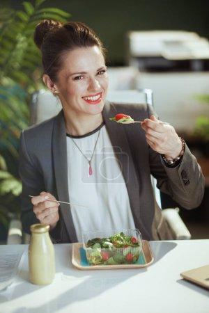 Photo for Sustainable workplace. happy modern middle aged woman worker in a grey business suit in modern green office eating salad. - Royalty Free Image