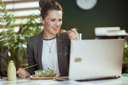 Téléchargez les photos : Un lieu de travail durable. travailleuse moderne souriante dans un costume d'affaires gris dans un bureau vert moderne avec salade mangeant ordinateur portable. - en image libre de droit