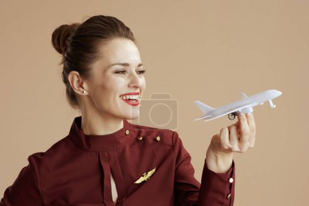 Photo for Smiling elegant female flight attendant against beige background with a little airplane. - Royalty Free Image