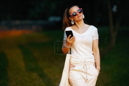 Téléchargez les photos : L'heure d'été. souriant élégante femme d'âge moyen en chemise blanche en utilisant smartphone et la marche. - en image libre de droit